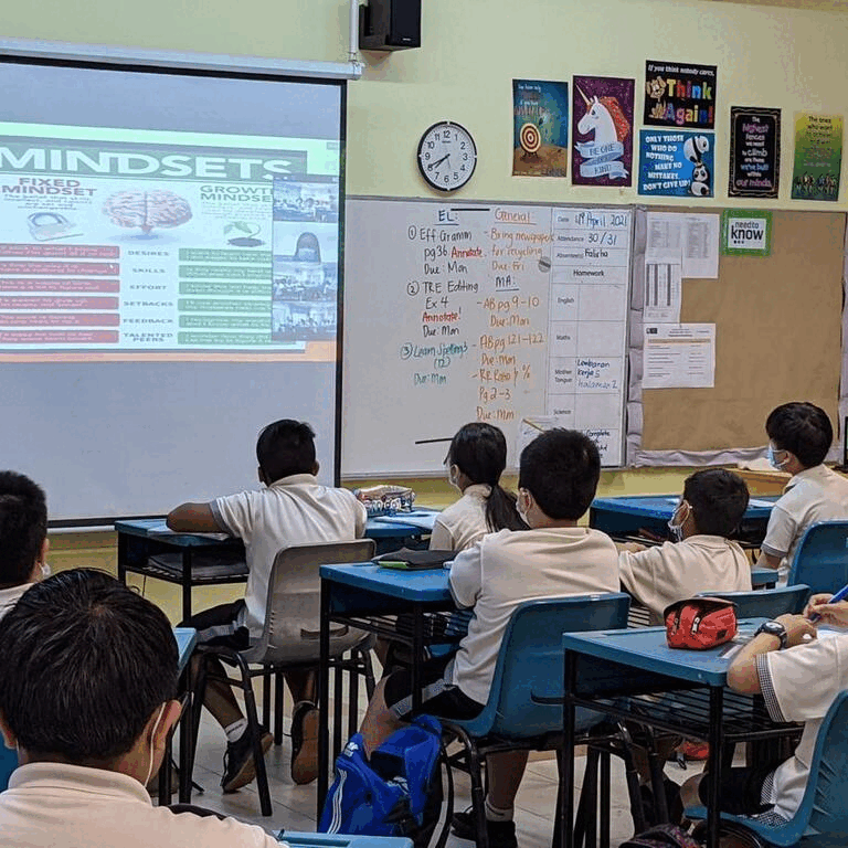 Students in uniforms sit at desks facing a projector slide titled Mindsets and a whiteboard with notes.